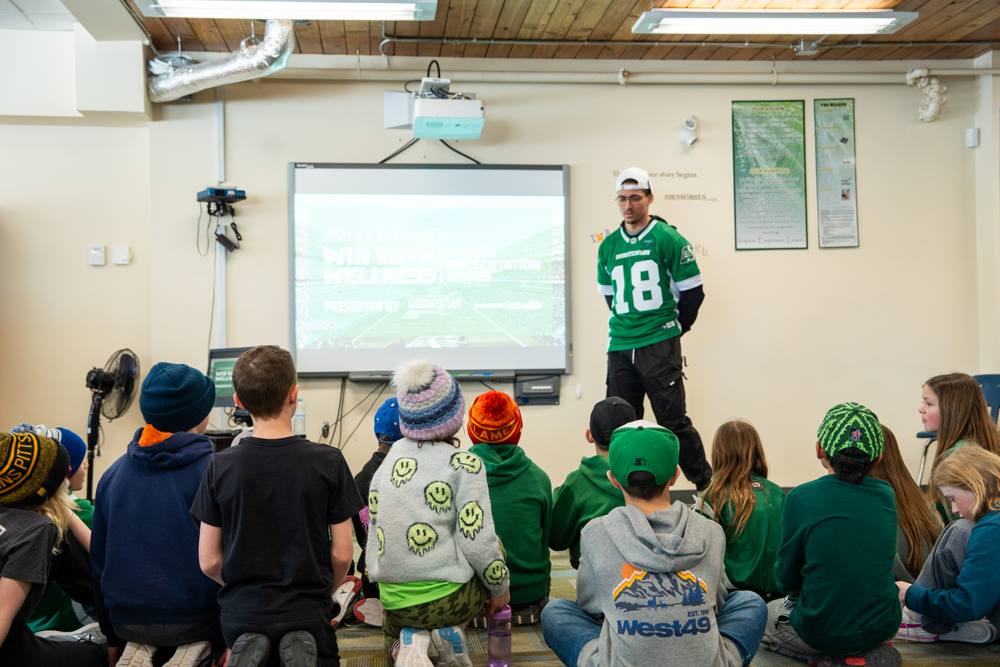 A Roughriders player presenting Win With Wellness to young students sitting on the floor