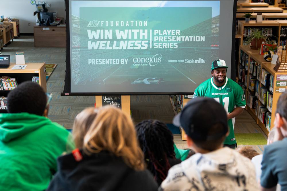 A smiling Roughriders player in front of the Win With Wellness screen in a school library