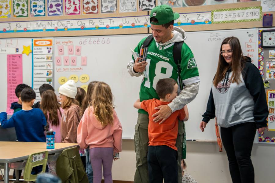 A Roughriders player hugging a young student in a classroom